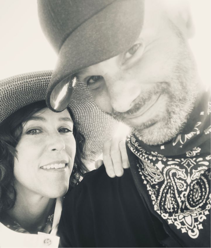 A close-up, black-and-white photo of two smiling adults wearing hats. The woman on the left wears a woven hat, and the man on the right wears a baseball cap and bandana around his neck. They appear happy and relaxed.